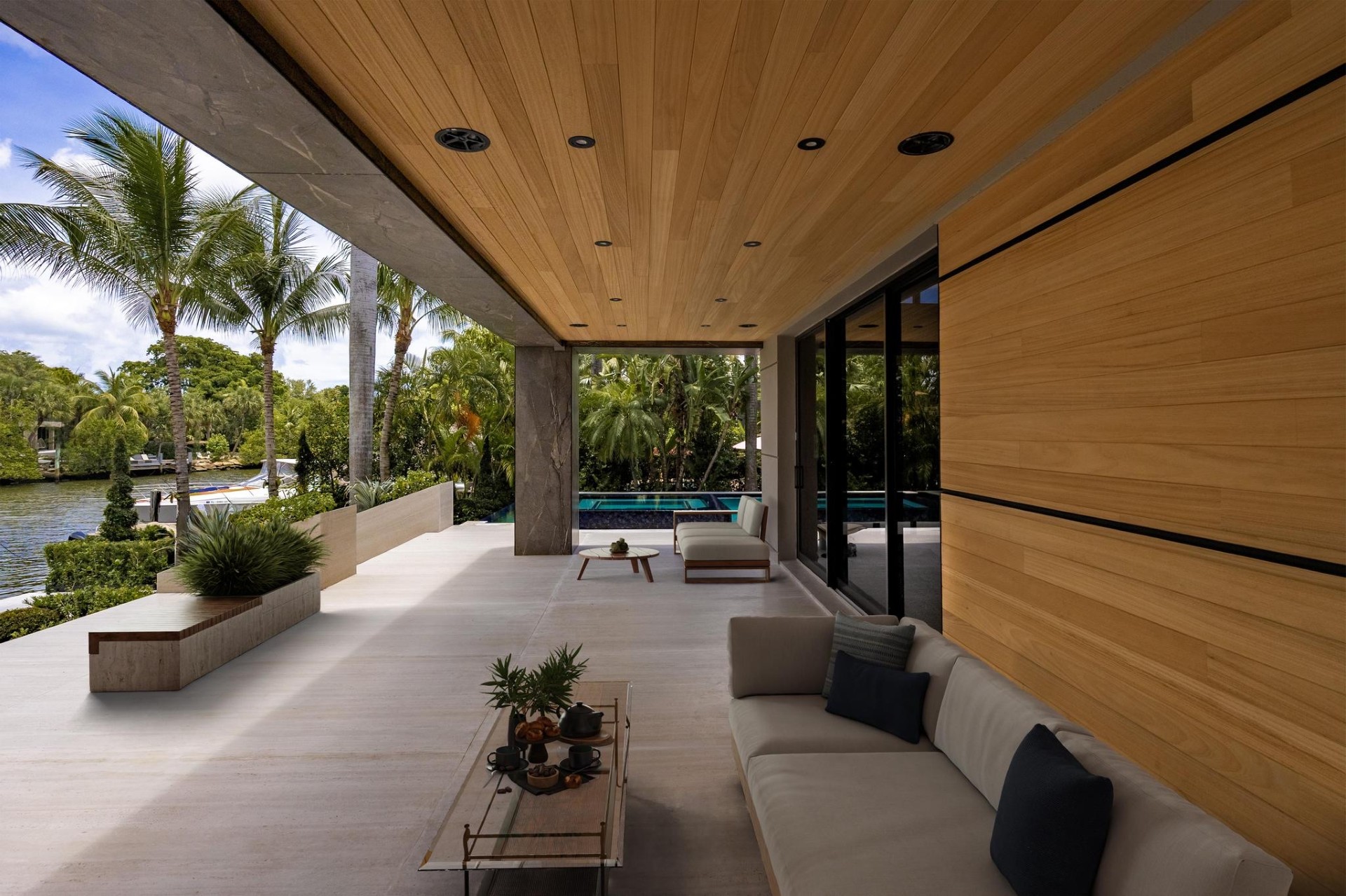 A covered outdoor patio with wood-paneled ceiling and walls, lounge seating, and a pool and palm trees in the background