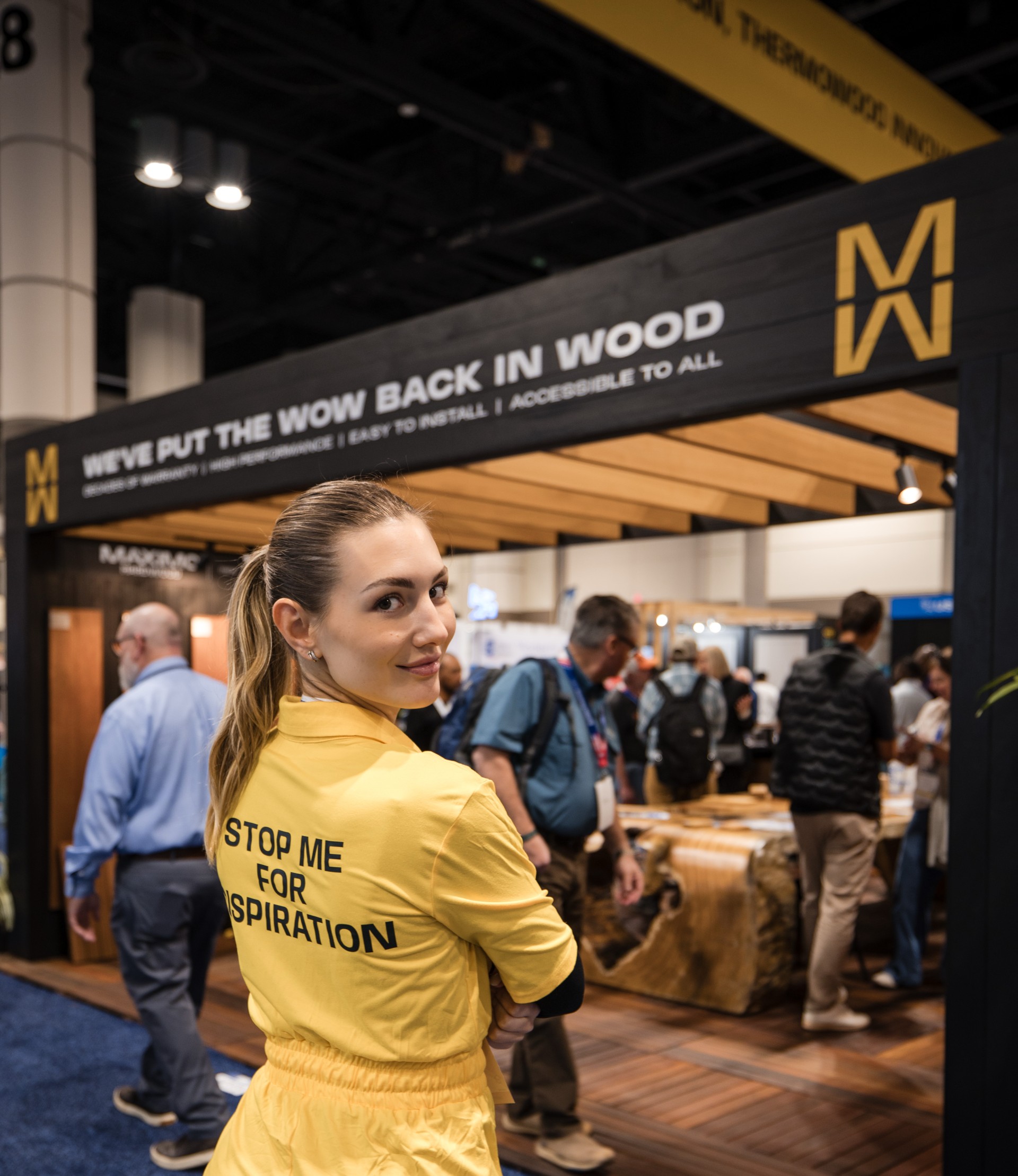 Ana Caovilla glancing back over her shoulder at a trade show booth, wearing a yellow jumpsuit reading "Stop me for inspiration," in front of the Maximo Wood display