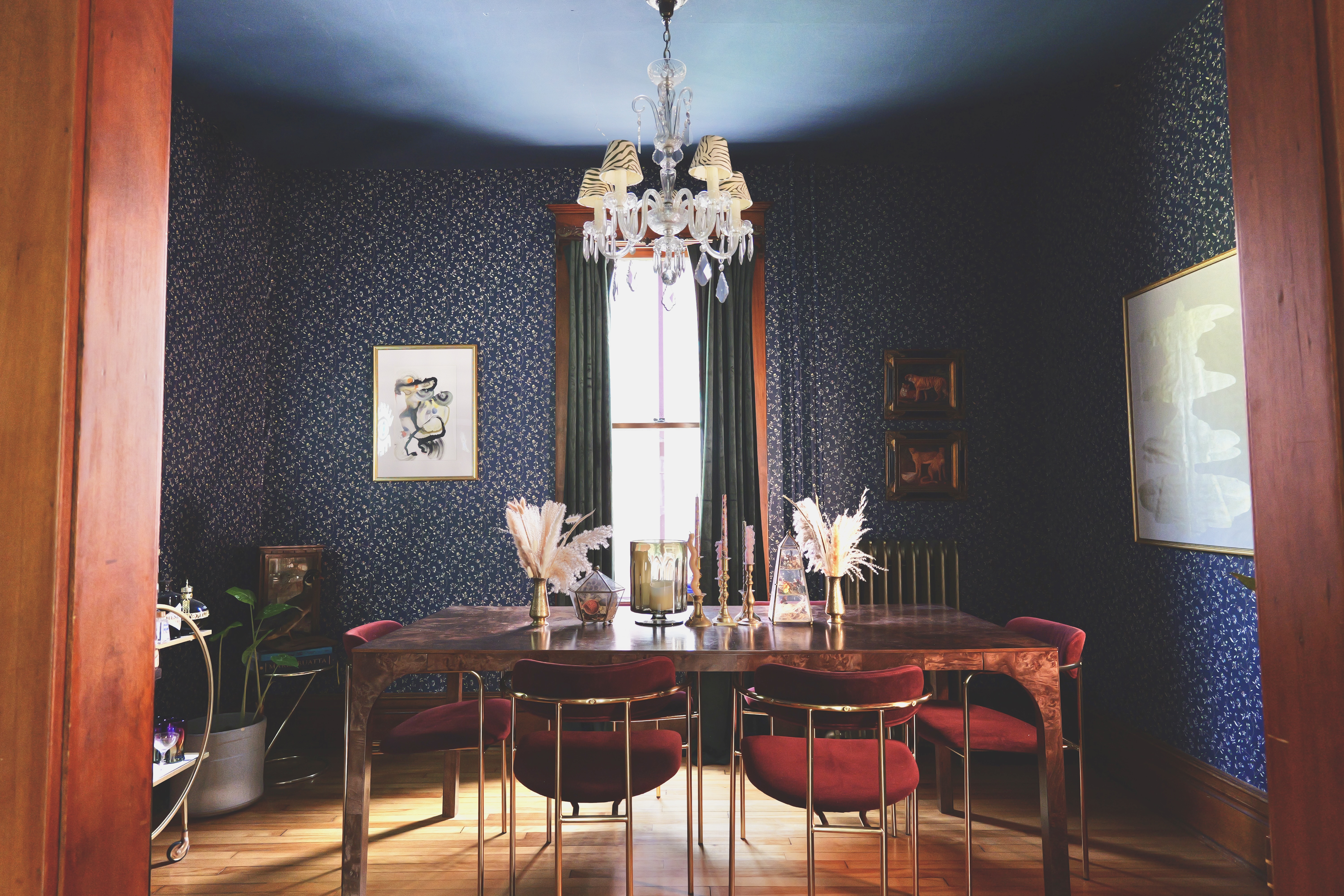 A dining room designed by Lauren Hunter Design, with dark floral wallpaper, a crystal chandelier, a burl-wood table, and red velvet chairs