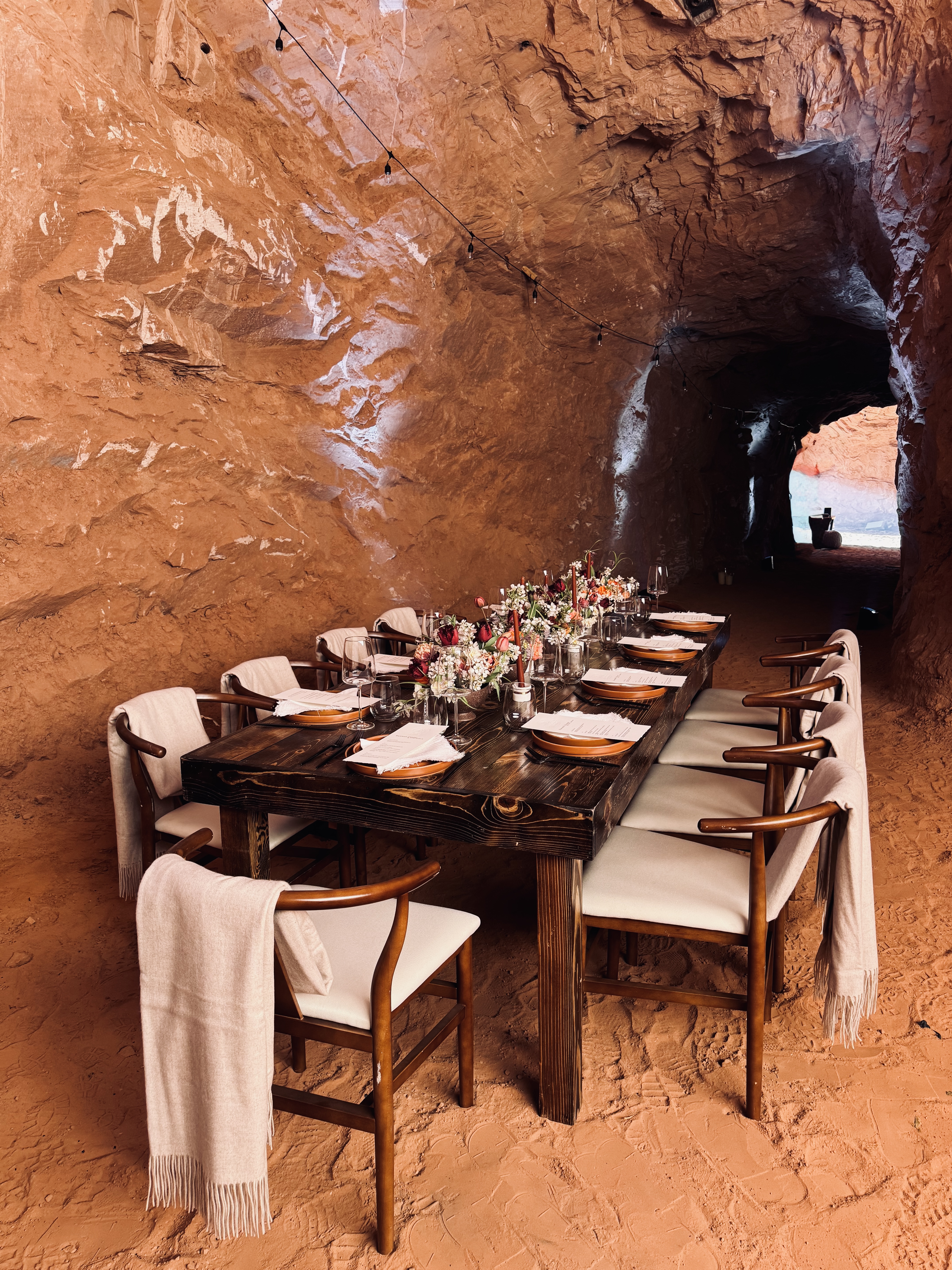 A long dinner table set for eight, draped chairs and string lights inside a sandstone cave with a tunnel opening to daylight beyond